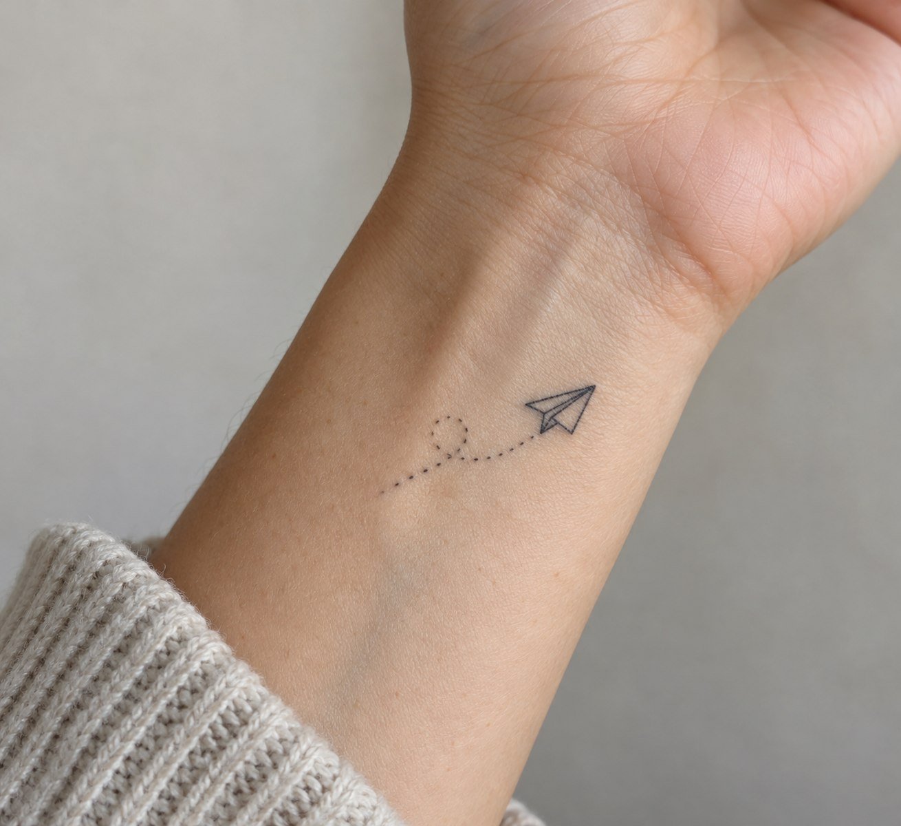 Minimal paper plane tattoo with dotted trail on the wrist.