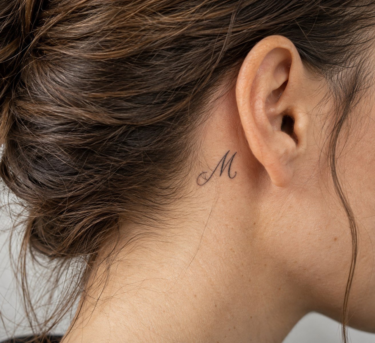 Single-letter tattoo behind the ear in fine script.